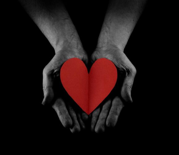 All black photo with two hands and a red heart in the middle.